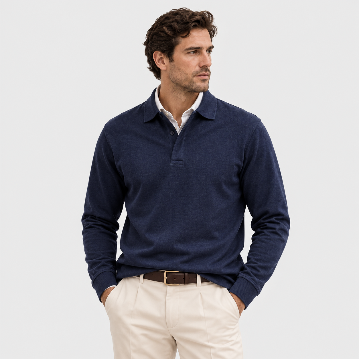 A man wearing a navy blue long-sleeve polo sweater over a white collared shirt, paired with beige trousers and a brown belt, photographed against a light gray studio background.