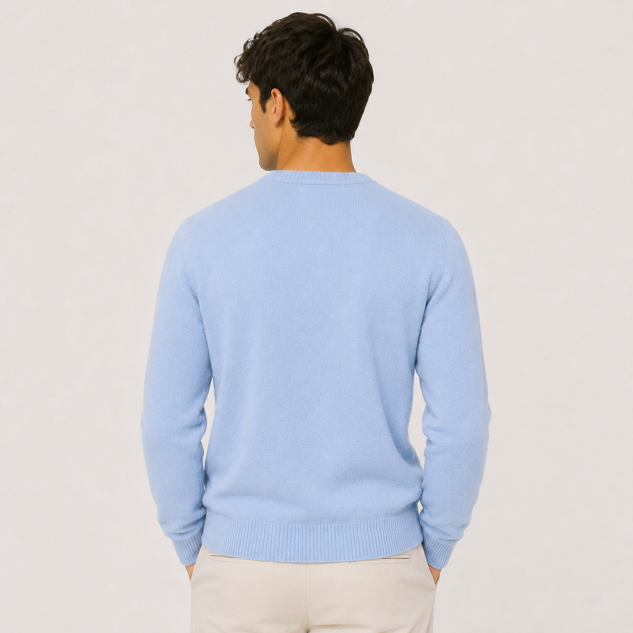 A man viewed from the back wearing a light blue crewneck sweater and cream trousers, standing against a plain light gray studio background.