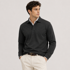 A man wearing a dark charcoal gray long-sleeve polo shirt over a white collared shirt, paired with cream-colored trousers and a brown belt, standing against a light gray studio background.