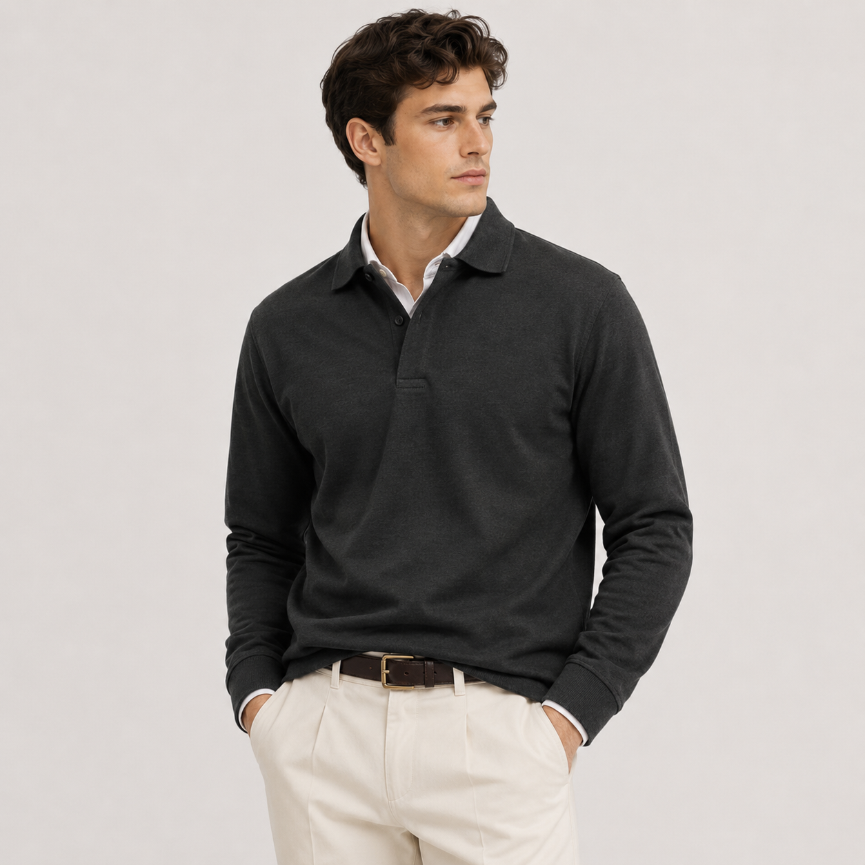 A man wearing a dark charcoal gray long-sleeve polo shirt over a white collared shirt, paired with cream-colored trousers and a brown belt, standing against a light gray studio background.