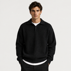 A man wearing a black knit polo sweater over a white undershirt, standing against a light gray studio background.