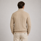 A man viewed from the back wearing a beige cable-knit mock-neck sweater and matching beige trousers, set against a plain light gray studio background.
