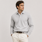 A man wearing a light gray long-sleeve polo sweater over a white collared shirt, paired with off-white trousers and a dark brown belt, photographed against a plain light gray studio background.