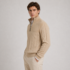 A man wearing a beige cable-knit quarter-zip sweater over a white collared shirt and beige trousers, standing against a light gray studio background.