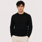 A man wearing a black crewneck knit sweater and off-white trousers, standing against a plain light gray studio background.
