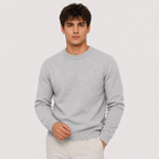 A man wearing a light gray crewneck knit sweater and off-white trousers, standing against a plain light gray studio background.