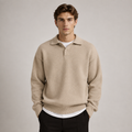 A man wearing a beige knit polo sweater over a white t-shirt and black trousers, standing against a neutral light gray background.