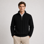 A man wearing a black quarter-zip knit sweater with cable and textured panel details, styled over a white collared shirt and beige trousers, against a light gray studio background.
