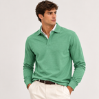 A man wearing a green long-sleeve polo shirt over a white collared shirt, paired with off-white trousers and a dark brown belt, photographed against a light gray studio background.