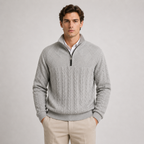 A man wearing a light gray cable-knit quarter-zip sweater over a white collared shirt and beige trousers, standing against a neutral light gray studio background.