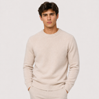A man wearing a light beige crewneck knit sweater and matching trousers, standing against a solid light gray background.