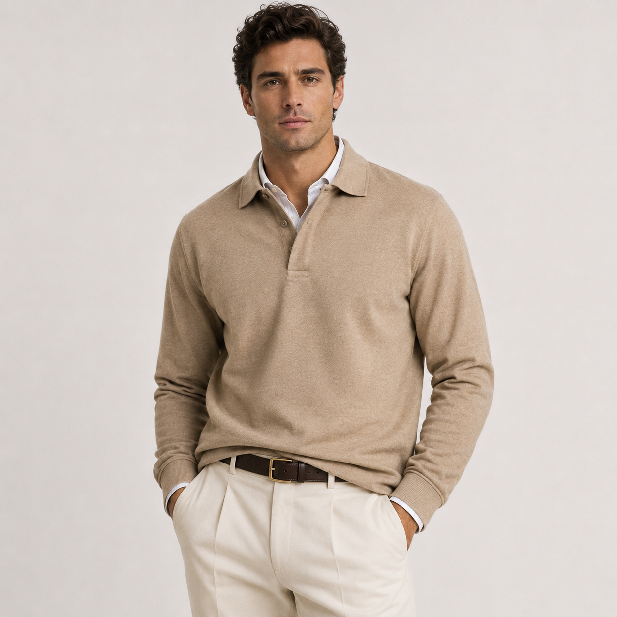 A man wearing a beige long-sleeve polo sweater over a white collared shirt, paired with cream trousers and a dark brown belt, standing against a light gray studio background.