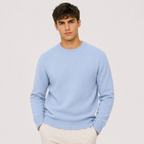 A man wearing a light blue crewneck knit sweater and cream trousers, standing against a plain light gray studio background.