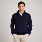 A man wearing a navy blue quarter-zip knit sweater with cable and rib textures, layered over a white collared shirt and paired with beige trousers, standing against a light gray studio background.
