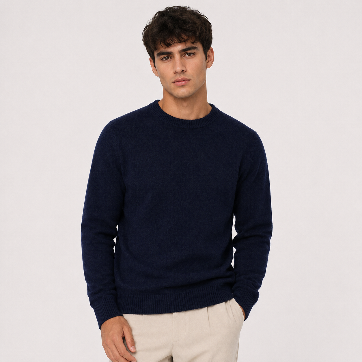 A man wearing a navy blue crewneck sweater and light beige trousers, standing against a plain light gray studio background.
