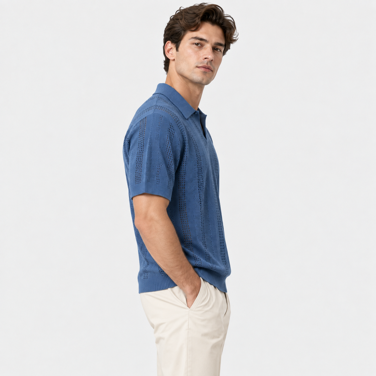 A man wearing a textured blue short-sleeve knit polo shirt and cream-colored trousers, standing in profile against a solid white studio background.