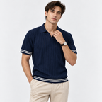 A man wearing a navy blue ribbed short-sleeve polo shirt with white stripes on the sleeves and hem, paired with beige trousers, against a light gray studio background.