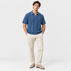 A man modeling a short-sleeve blue button-up shirt with vertical textured stripes, paired with beige trousers and beige loafers, against a light gray studio background.