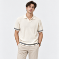 A man wearing a cream-colored ribbed short-sleeve polo shirt with black and white striped trim on the collar, cuffs, and hem, paired with beige trousers, against a light gray studio background.