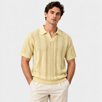 A man wearing a pale yellow knit short-sleeve polo shirt with a textured openwork pattern, paired with cream trousers, against a light gray studio background.