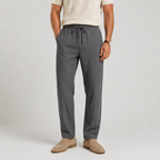 A man wearing dark gray drawstring linen trousers, paired with a cream t-shirt and beige loafers, standing against a light gray studio background.
