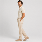 A man wearing a light beige short-sleeve polo shirt, matching beige drawstring trousers, and beige loafers, standing in profile against a light gray studio background.