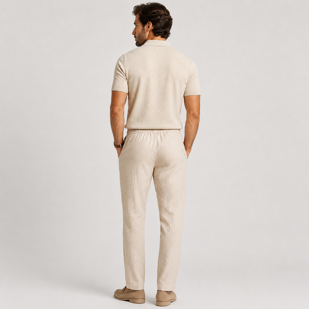 A man viewed from behind wearing a light beige short-sleeve knit polo shirt and matching beige trousers, paired with tan loafers, against a light gray studio background.