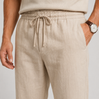 Beige linen drawstring trousers shown on a model, featuring an elastic waistband, side pockets, and a relaxed fit.
