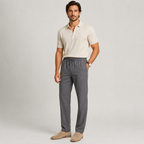 A man modeling a light beige short-sleeve polo shirt and dark gray drawstring trousers, paired with beige loafers, against a light gray studio background.