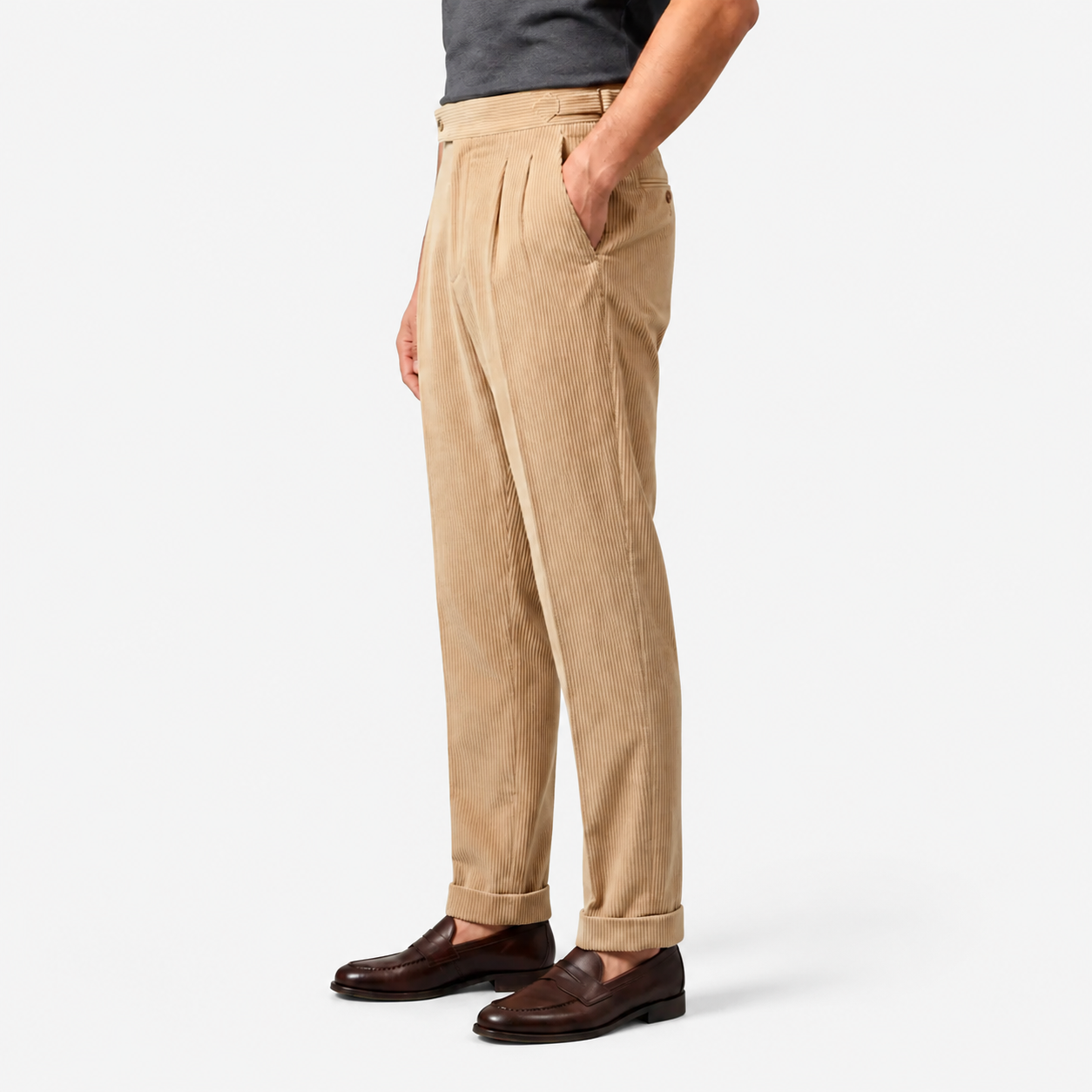 A person wearing beige corduroy trousers with a dark gray shirt and dark brown leather loafers, shown from the waist down against a light gray studio background.