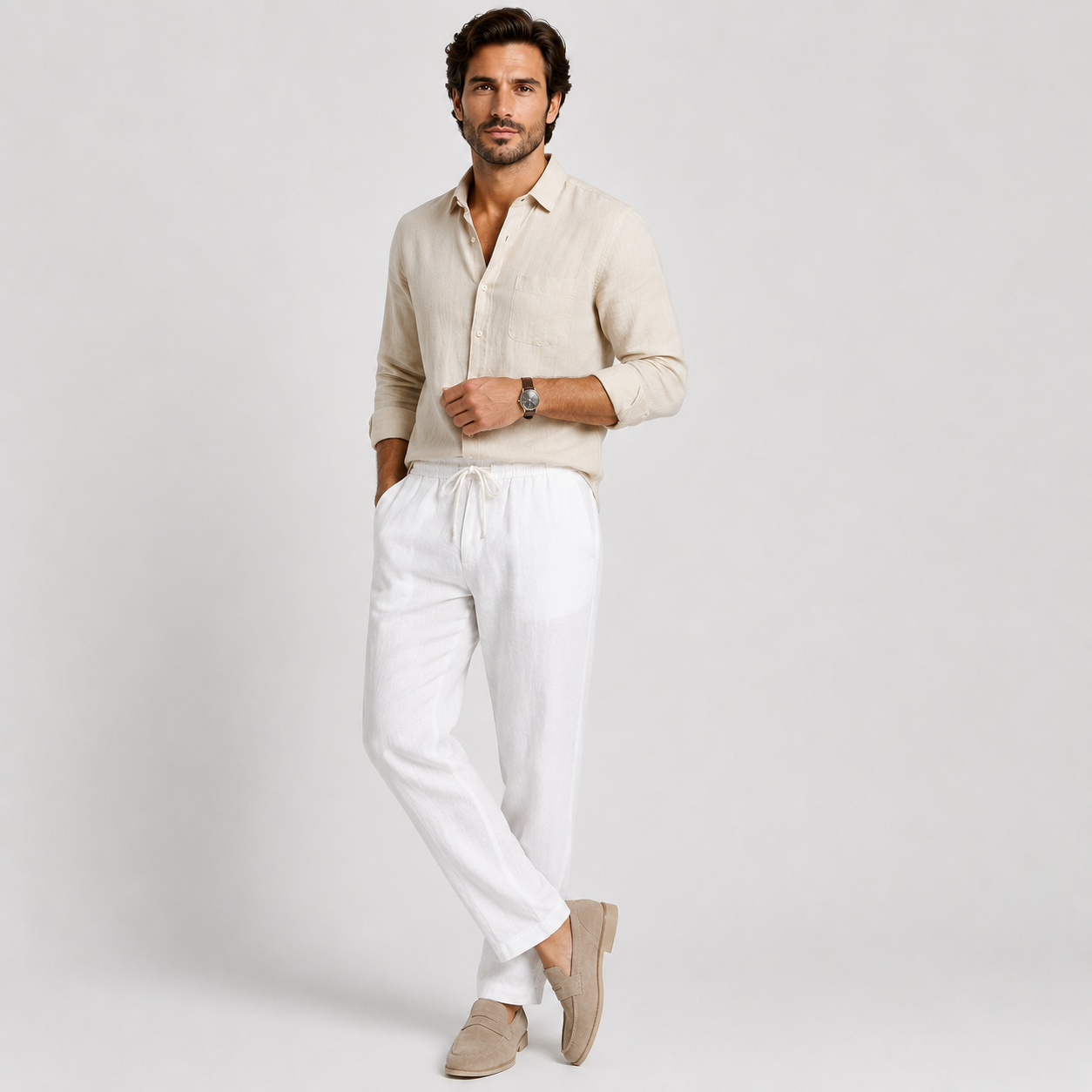 A man modeling a light beige linen button-down shirt and white linen drawstring trousers, paired with beige loafers, against a light gray studio background.
