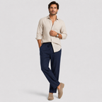 A man modeling a light beige linen button-down shirt and navy blue drawstring trousers, paired with beige loafers, against a light gray studio background.