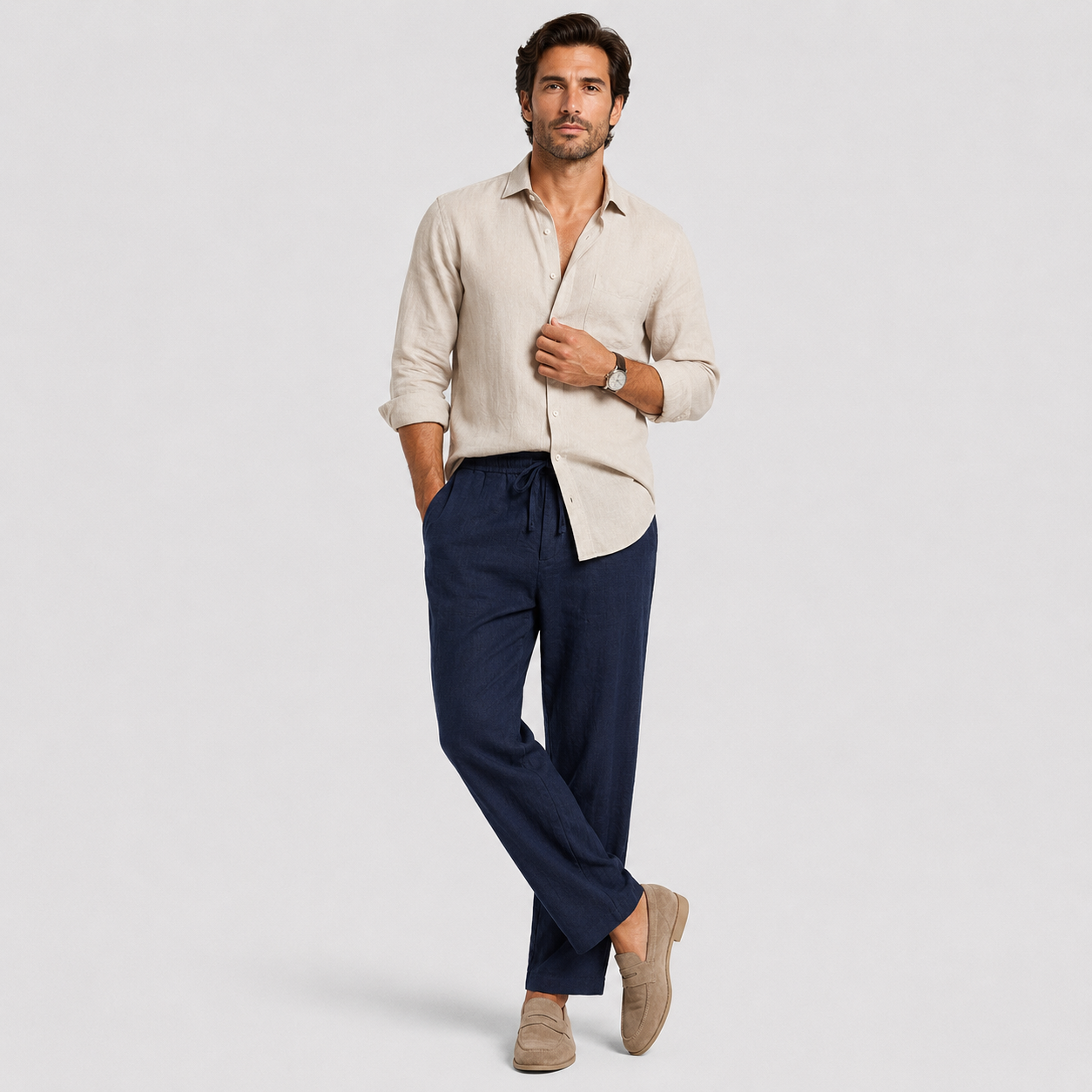 A man modeling a light beige linen button-down shirt and navy blue drawstring trousers, paired with beige loafers, against a light gray studio background.