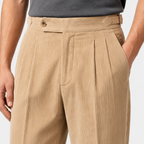 Close-up of a person wearing tan corduroy trousers with pleats, a matching belt, and a dark gray shirt against a light gray studio background.