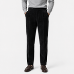 A person wearing black corduroy trousers with a gray sweater and brown leather shoes, standing against a light gray studio background.