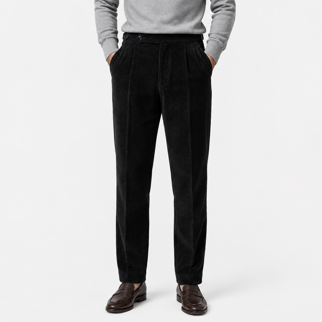 A person wearing black corduroy trousers with a gray sweater and brown leather shoes, standing against a light gray studio background.