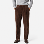 A person wearing dark brown corduroy trousers with a gray long-sleeve top and dark brown leather dress shoes, shown from the waist down on a light gray studio background.