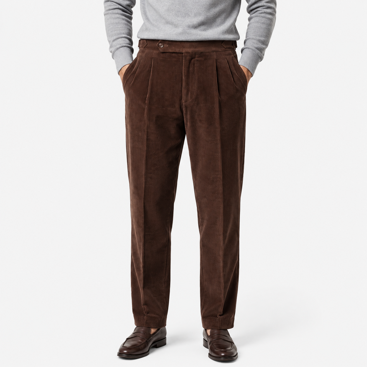 A person wearing dark brown corduroy trousers with a gray long-sleeve top and dark brown leather dress shoes, shown from the waist down on a light gray studio background.