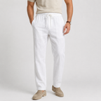 A man wearing white drawstring linen pants, a beige t-shirt, and beige loafers, standing against a light gray studio background.