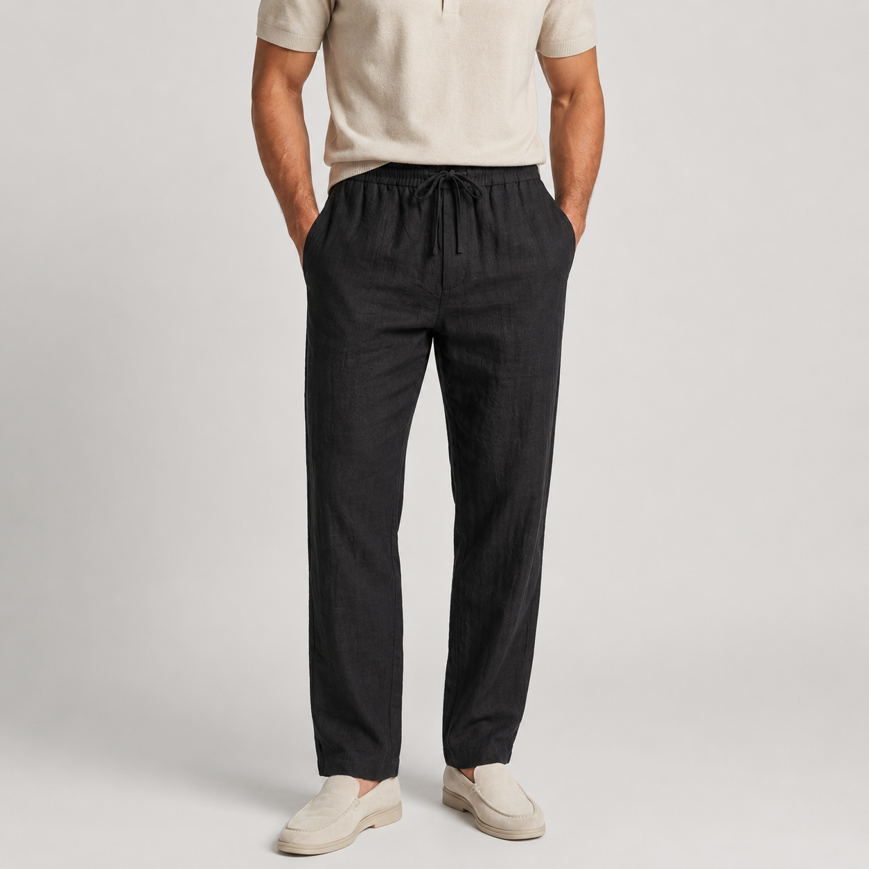 A man wearing dark charcoal gray drawstring trousers, a light beige short-sleeve polo shirt, and beige loafers, standing against a light gray studio background.