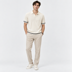 A man modeling a cream ribbed short-sleeve polo shirt with black and white striped trim on the collar, cuffs, and hem, paired with beige trousers and white sneakers, against a light gray studio background.