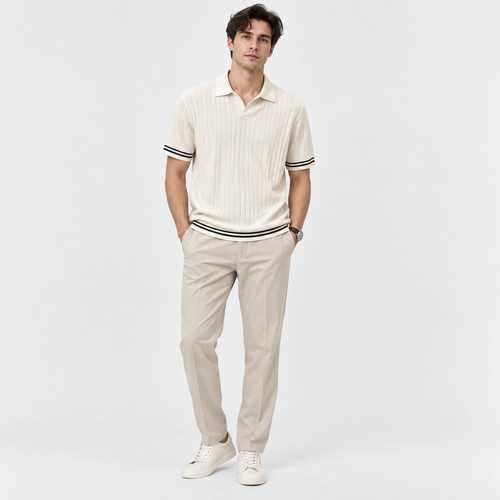 A man modeling a cream ribbed short-sleeve polo shirt with black and white striped trim on the collar, cuffs, and hem, paired with beige trousers and white sneakers, against a light gray studio background.