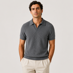 A man wearing a dark gray ribbed short-sleeve polo shirt and light beige trousers, standing against a light gray studio background.