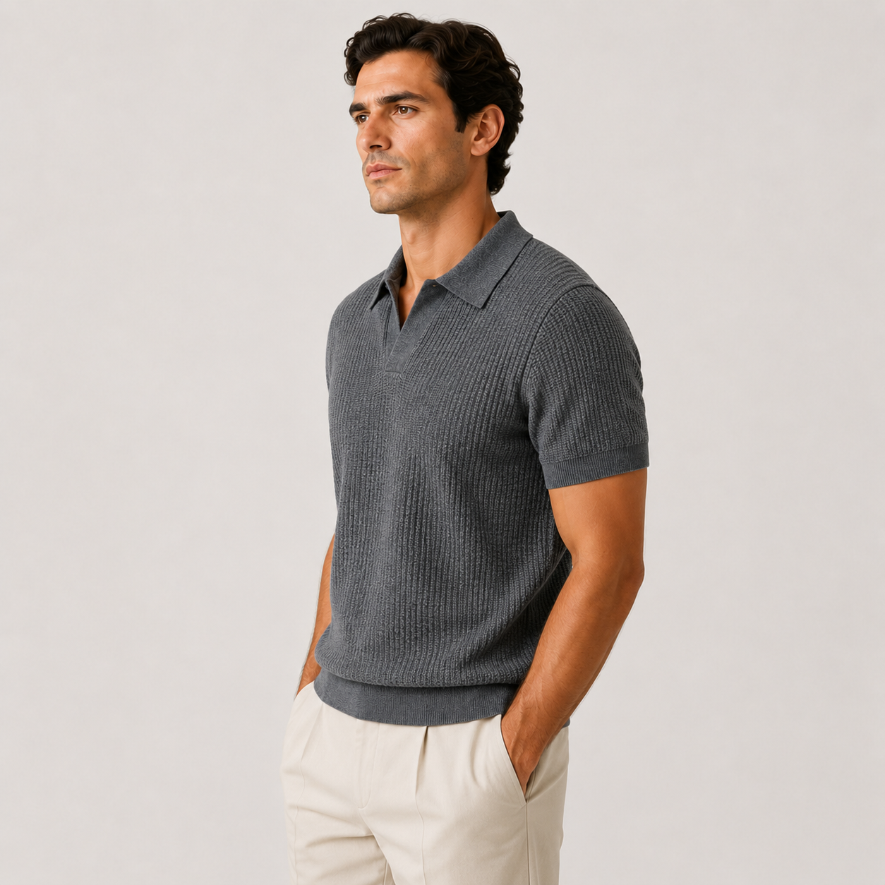 A man wearing a dark gray ribbed short-sleeve polo shirt and off-white trousers, standing against a light gray studio background.