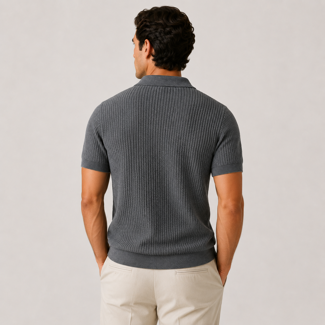 A man viewed from behind wearing a dark gray ribbed short-sleeve polo shirt and light beige trousers, standing against a plain light gray studio background.