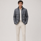 A man modeling a dark gray puffer jacket over a white shirt and cream trousers, standing against a light gray studio background.
