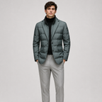 A man modeling a dark slate green puffer blazer over a black turtleneck sweater and light gray trousers, standing against a solid light gray studio background.