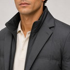 Close-up of a man wearing a dark charcoal gray puffer jacket with a high collar over a cream-colored collared shirt. The jacket has subtle circular quilted details and a tonal logo on the chest pocket.