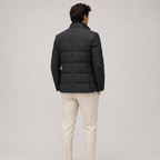 A man viewed from behind wearing a black puffer jacket and light beige trousers, standing against a neutral light gray studio background.