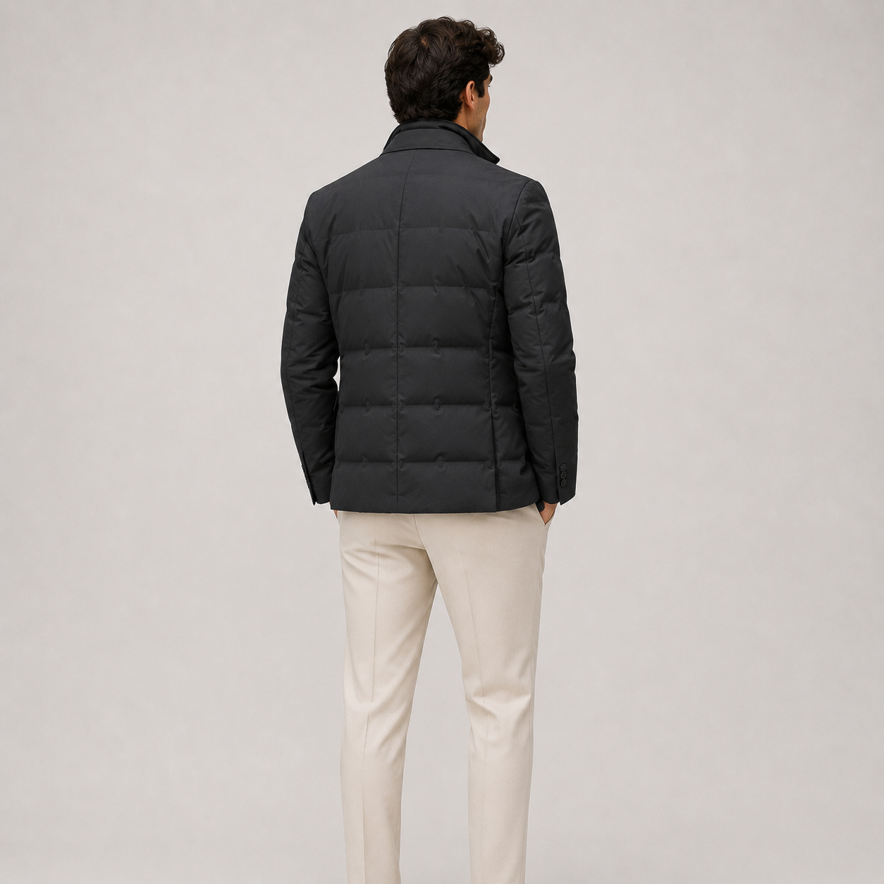 A man viewed from behind wearing a black puffer jacket and light beige trousers, standing against a neutral light gray studio background.